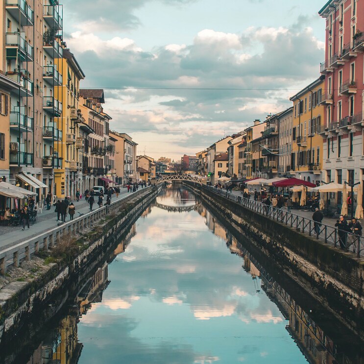 A scenic view of the Navigli canal showcases charming buildings lining the waterway. Reflections of clouds and structures create a serene atmosphere. People stroll along the banks, while outdoor dining spaces are visible under colorful awnings.