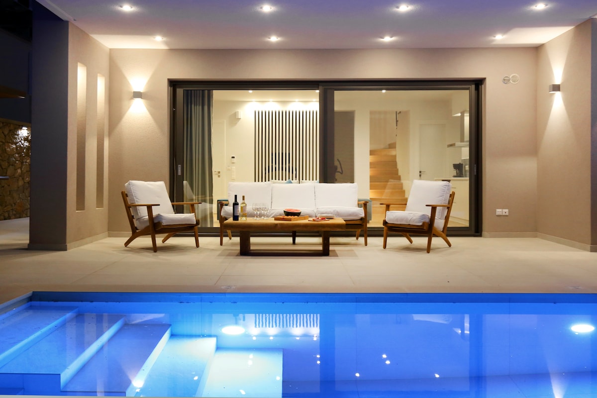 An outdoor lounge area features a wooden table surrounded by comfortable seating, positioned next to a reflective blue swimming pool. Large sliding glass doors provide a view into the interior space, flanked by soft ambient lighting that highlights the structure's modern design.