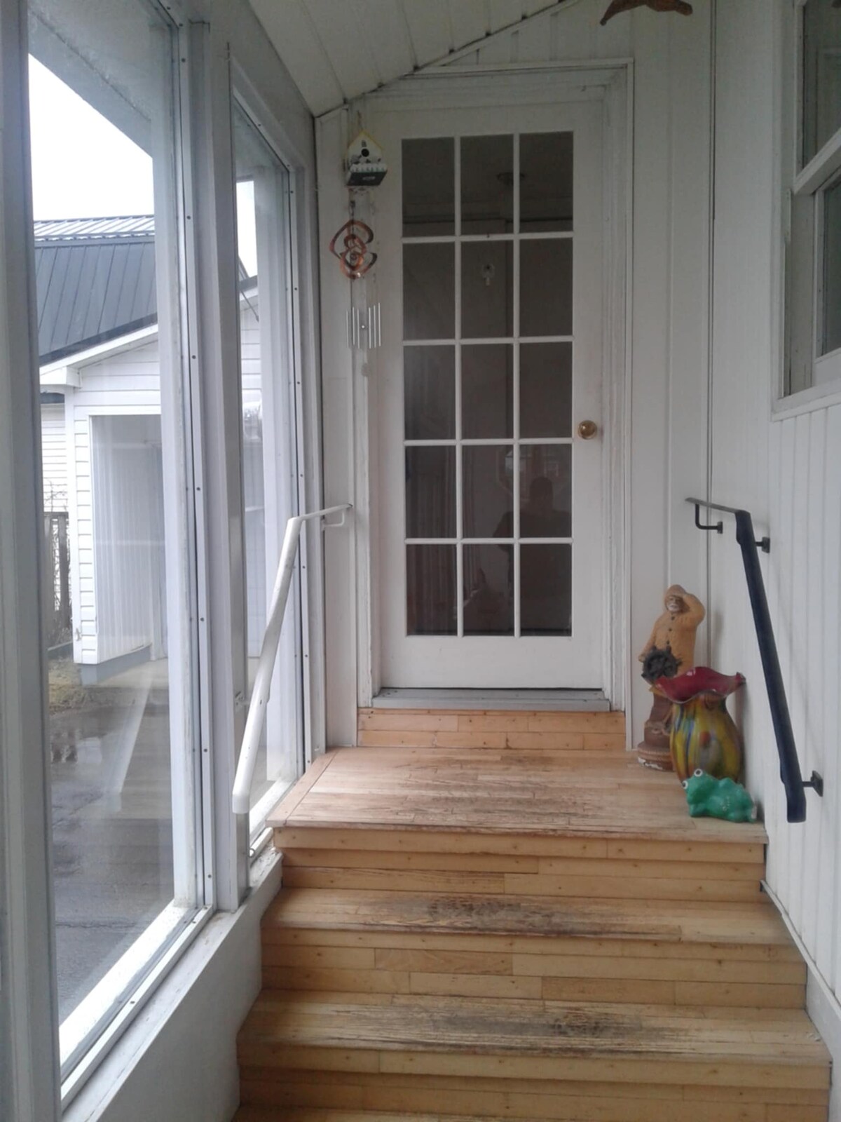 A welcoming entrance is captured, featuring a wooden staircase leading up to a glass-paneled door. Natural light filters through large windows on either side, while decorative items, including a colorful statue, are positioned at the base of the steps.