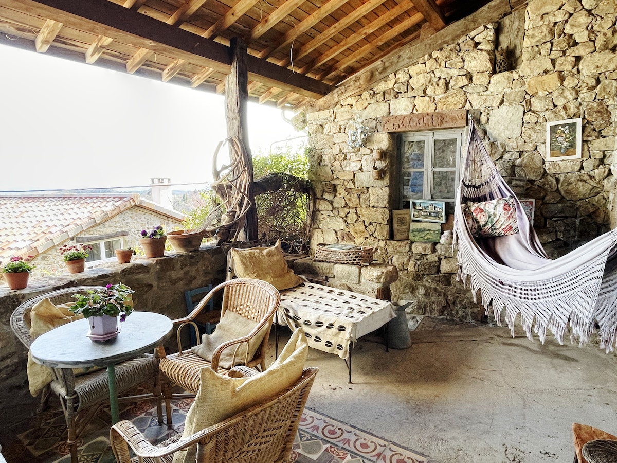 A cozy patio area is showcased with natural stone walls and a wooden ceiling. Comfortable seating options, including a hammock and woven chairs, surround a small table. Potted plants add greenery, while a patterned floor enhances the rustic charm of the space.