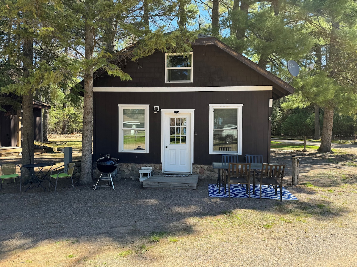 A charming cabin exterior is showcased, featuring natural wood siding and a white door. A small outdoor seating area is set up with a patterned rug and black chairs. Nearby, a charcoal grill rests beside trees providing shade.