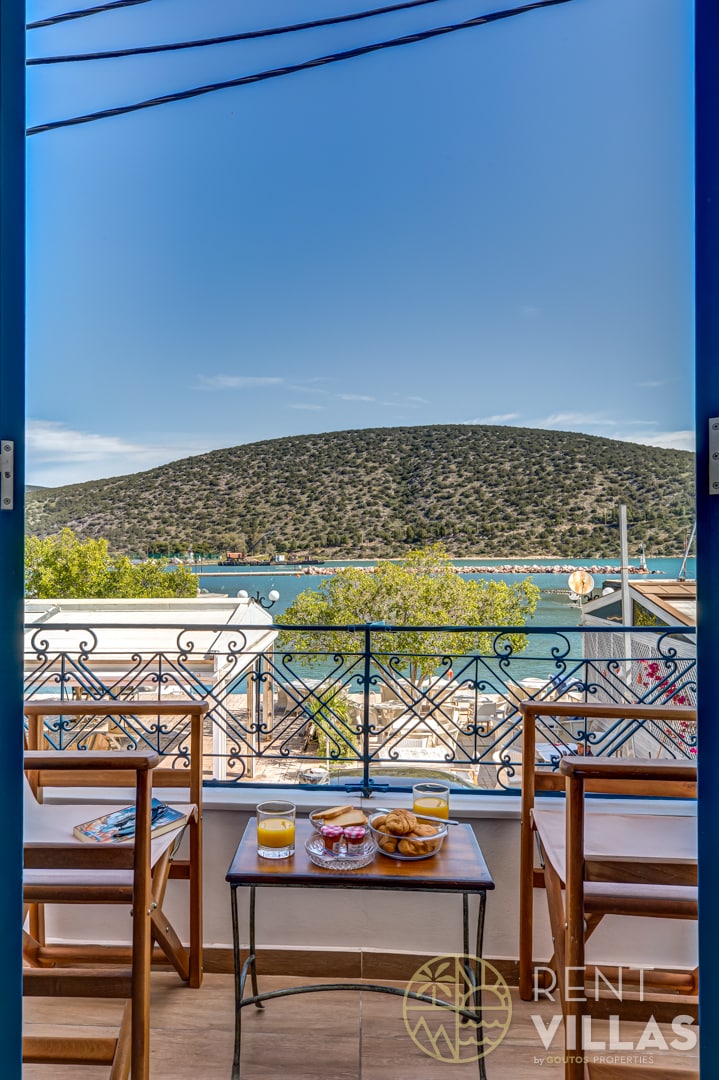 A view from the balcony showcases a serene landscape with a mountain in the distance and a calm sea below. A small table with refreshments is visible, complemented by two chairs, inviting relaxation in the beautiful outdoor setting.