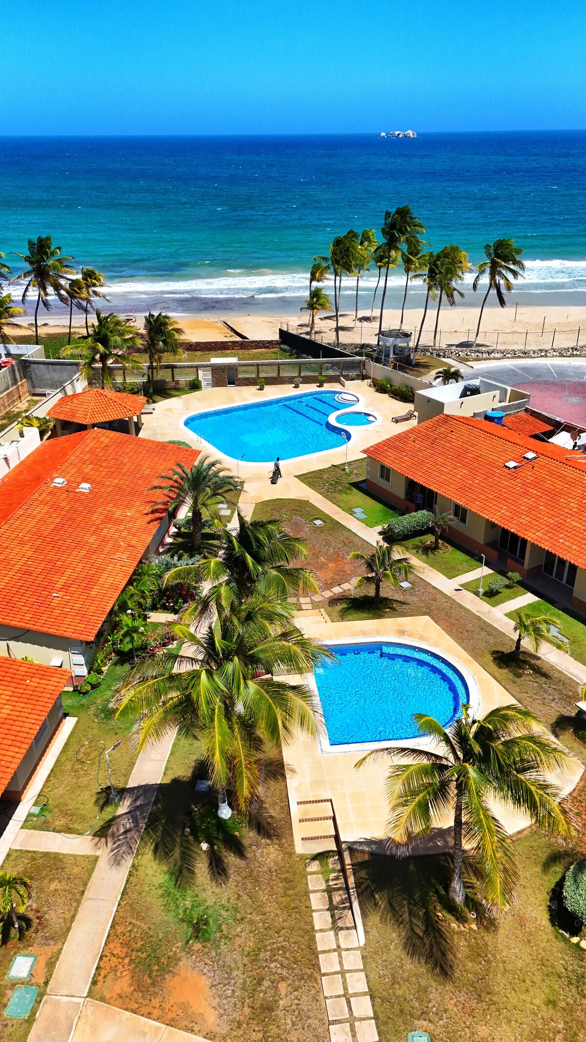 An aerial view reveals two swimming pools surrounded by well-maintained green lawns and palm trees. The beach is visible in the background, with a clear ocean meeting a bright sky. Colorful rooftops of nearby buildings create a vibrant contrast with the natural scenery.