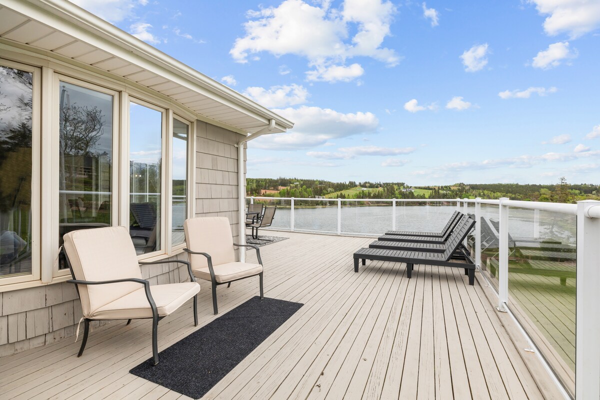 A spacious deck extends towards the waterfront, furnished with two lounge chairs and a small accent rug. Expansive windows provide views of the surrounding landscape while glass railings ensure safety and transparency, allowing sunlight to enhance the serene atmosphere.
