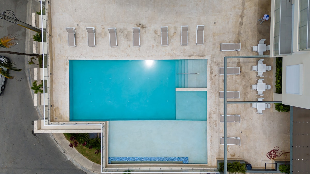 An aerial view captures the swimming pool, featuring a clear blue surface and surrounding lounge chairs. A shallow area is visible at one end, and adjacent seating areas are arranged thoughtfully, enhancing the outdoor space for relaxation.