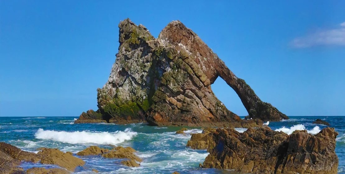 A natural rock formation stands prominently in the ocean, featuring a distinct arch that has formed over time. Waves crash against the rocky shoreline, creating a dynamic coastal scene under a clear blue sky.