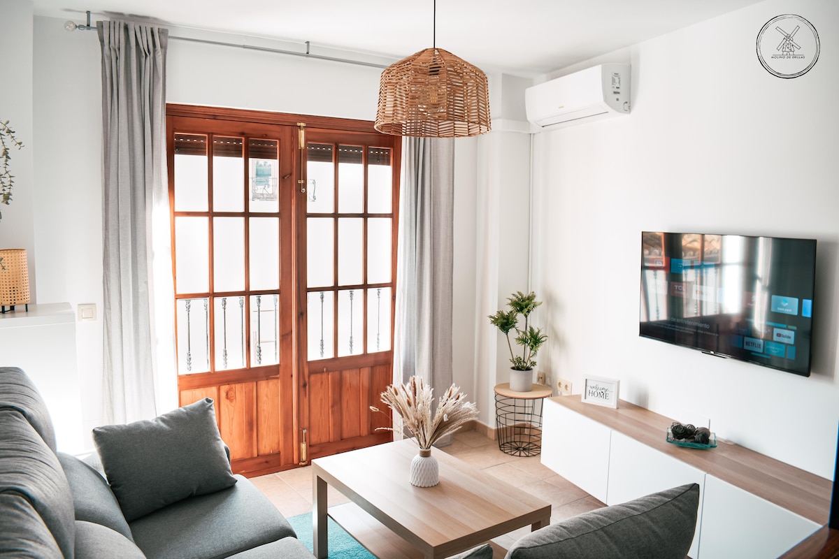A cozy living room is presented, featuring a comfortable gray sofa and a wooden coffee table. Natural light filters through double doors with wooden frames, leading to a small balcony. An air conditioning unit is visible, along with a potted plant and a flat-screen TV mounted on the wall.