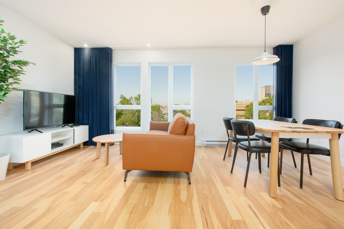 A modern living space features an inviting brown sofa facing a flat-screen television. A wooden dining table with black chairs is positioned next to large windows that offer unobstructed views of the outside. Bright wooden flooring complements the room's contemporary design.