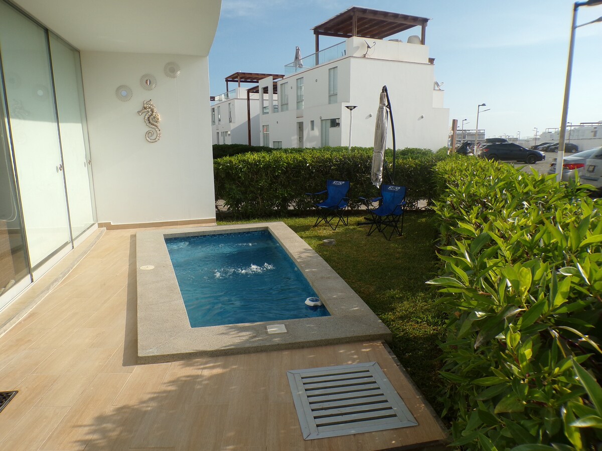 A small private plunge pool is positioned next to a grassy area, surrounded by low greenery. Two blue chairs are arranged nearby. The exterior features a light-colored building and clear sky in the background, with gentle ripples visible in the water.