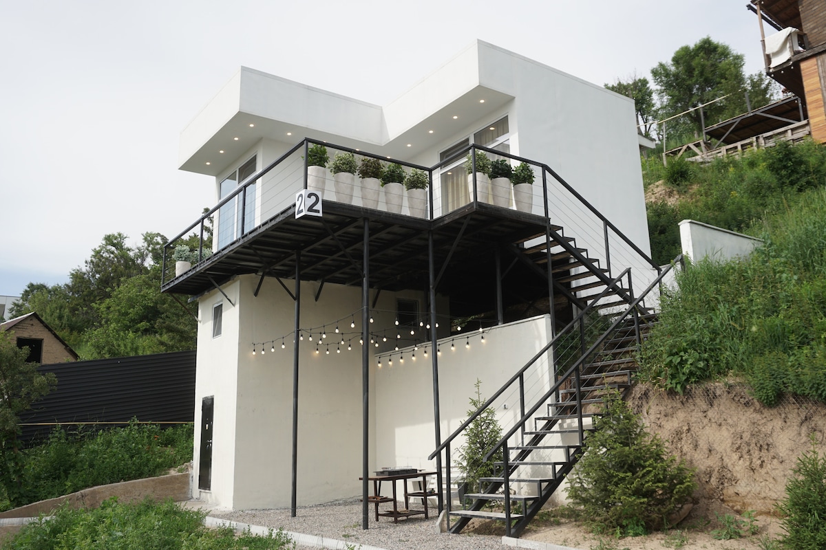 A modern house is showcased from an outdoor perspective, featuring a sleek white exterior and large windows. A staircase leads to an upper level, adorned with potted plants. Decorative lights are strung across the outdoor seating area, creating a welcoming environment.