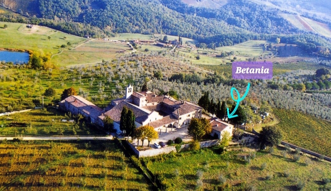 An aerial view of Betania showcases the historic structure nestled among expansive olive groves and vineyards. Rolling hills create a serene backdrop, while a clear blue sky complements the lush green landscape. The charm of the Tuscan countryside is visibly highlighted.