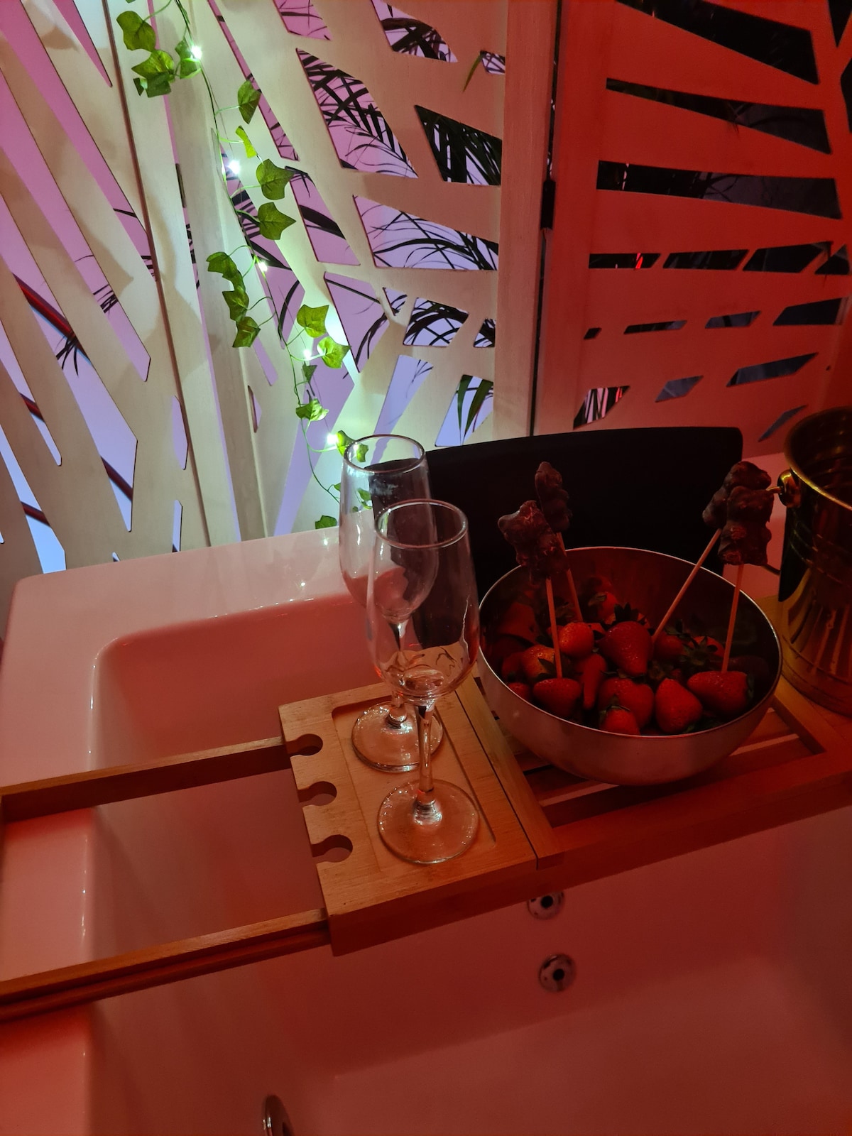 A wooden tray rests across the edge of a spa bathtub, featuring two champagne flutes and a bowl of strawberries. Soft lighting accentuates the surroundings, while a decorative screen with greenery adds privacy and ambiance.