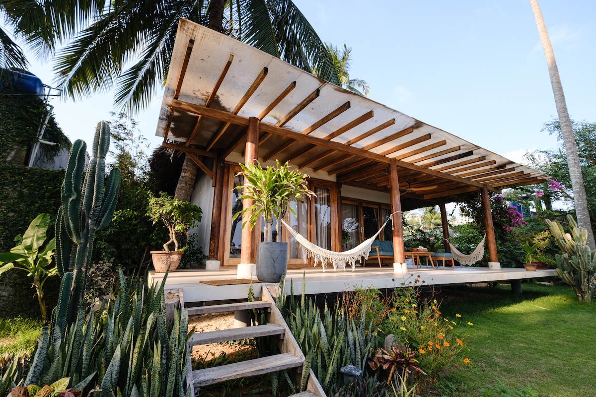 The beachfront villa features a spacious elevated porch with hammocks and seating arrangements. Lush greenery surrounds the property, complemented by tropical plants and decorative cacti. The structure showcases a traditional open design with wooden beams and large glass doors, allowing for natural light.