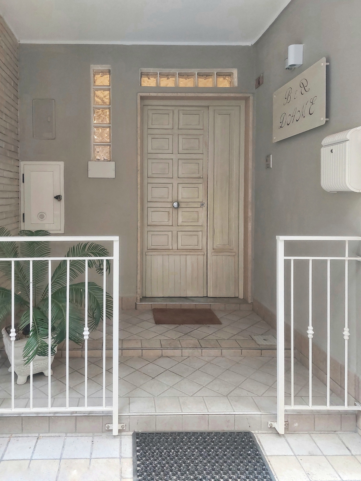 The entrance features a light-colored wooden door, flanked by a textured wall. A potted plant adds a touch of greenery near the tiled floor, while a welcome mat provides a hospitable touch. An intercom and mailboxes are visible along the side wall.