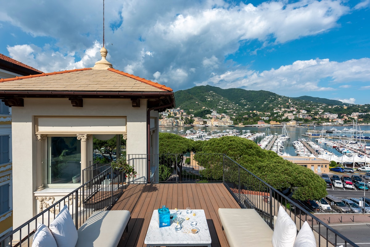 A rooftop terrace overlooks the marina and green hills of Portofino. Comfortable seating and a coffee table are arranged on a wooden deck, inviting relaxation. Scenic views of the surrounding landscape enhance the tranquil setting, with clouds casting soft shadows over the water.
