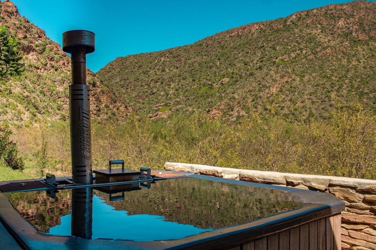 A wood-fired hot tub is positioned in the foreground, reflecting the clear blue sky. Surrounding mountains and greenery create a tranquil backdrop, enhancing the inviting nature of the outdoor space.