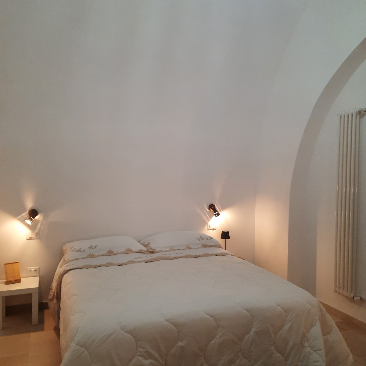 A spacious bedroom features a large bed with a light-colored quilt. Wall-mounted lamps provide soft illumination, and a small bedside table is positioned next to the bed. The room is characterized by arched ceilings and minimalist decor, offering a calm and serene atmosphere.