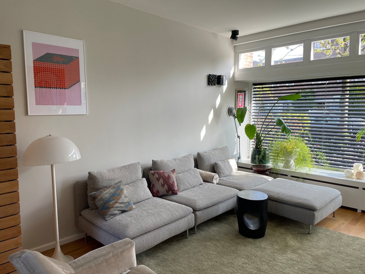 A light-filled living room is presented, featuring a spacious sectional couch with decorative cushions. A small black coffee table is positioned in front, while large windows allow sunlight to illuminate the room. A few potted plants add greenery to the serene atmosphere.