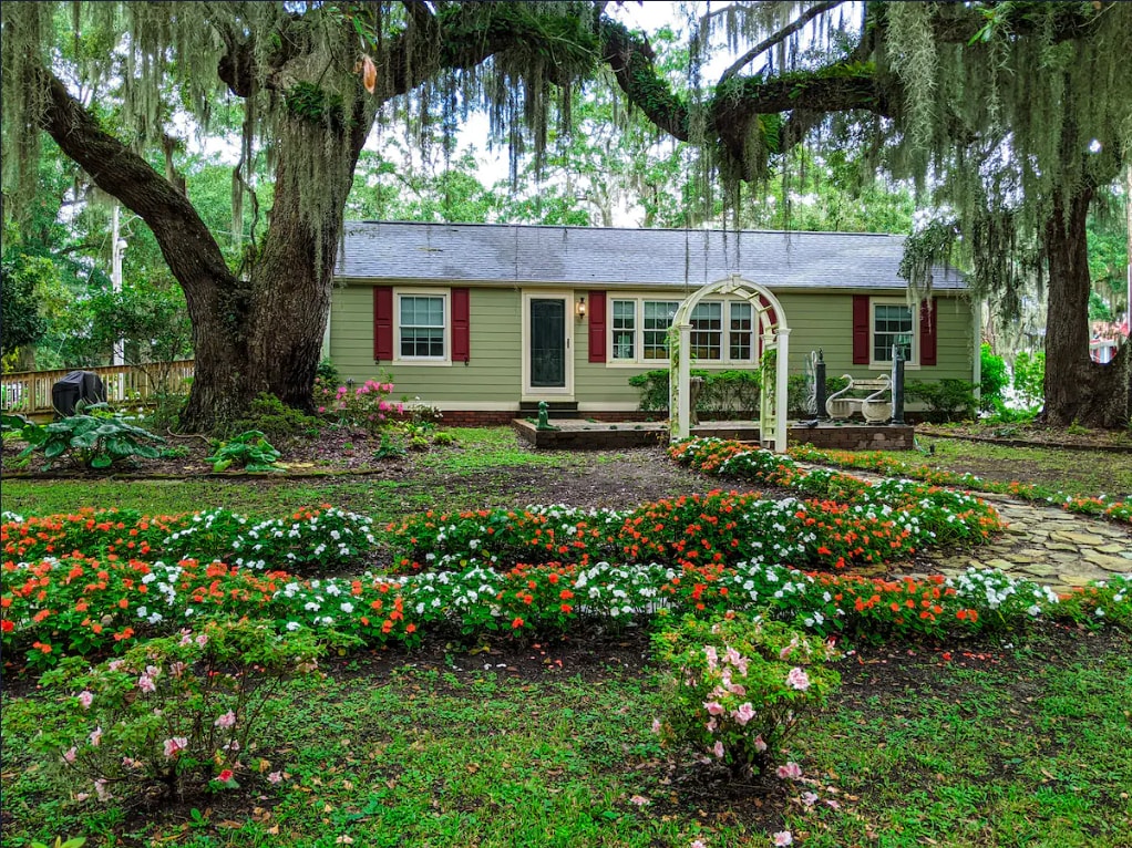 A charming cottage is surrounded by lush greenery and vibrant flower beds. Large trees provide shade, and a decorative archway leads to the entrance. The well-maintained garden features a mix of colorful blooms and manicured shrubs, creating a serene outdoor environment.