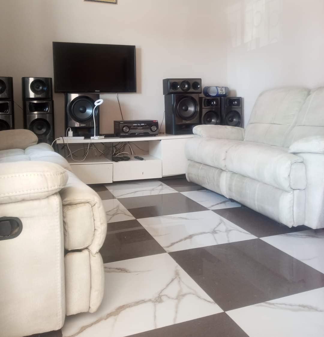 A cozy living area is highlighted by two light-colored recliners positioned around a modern entertainment unit. Speakers line the walls, and a flat-screen television rests atop the unit, with a sleek media player visible underneath. A marble-patterned floor adds a stylish touch.