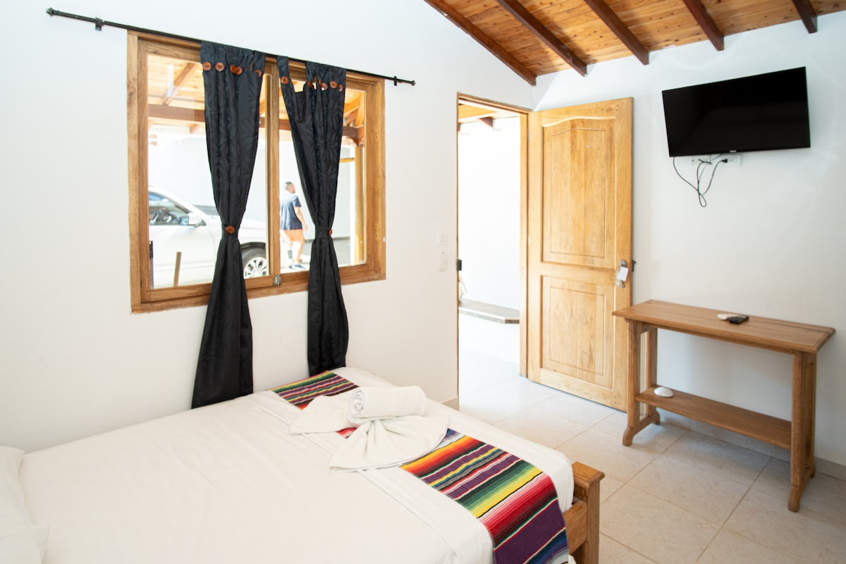 A sleeping area includes a spacious bed covered with a striped blanket, accompanied by white linens. Dark curtains frame the window, while wooden accents add warmth. A flat-screen TV is mounted on the wall above a simple wooden table, and a door leads to a hallway.