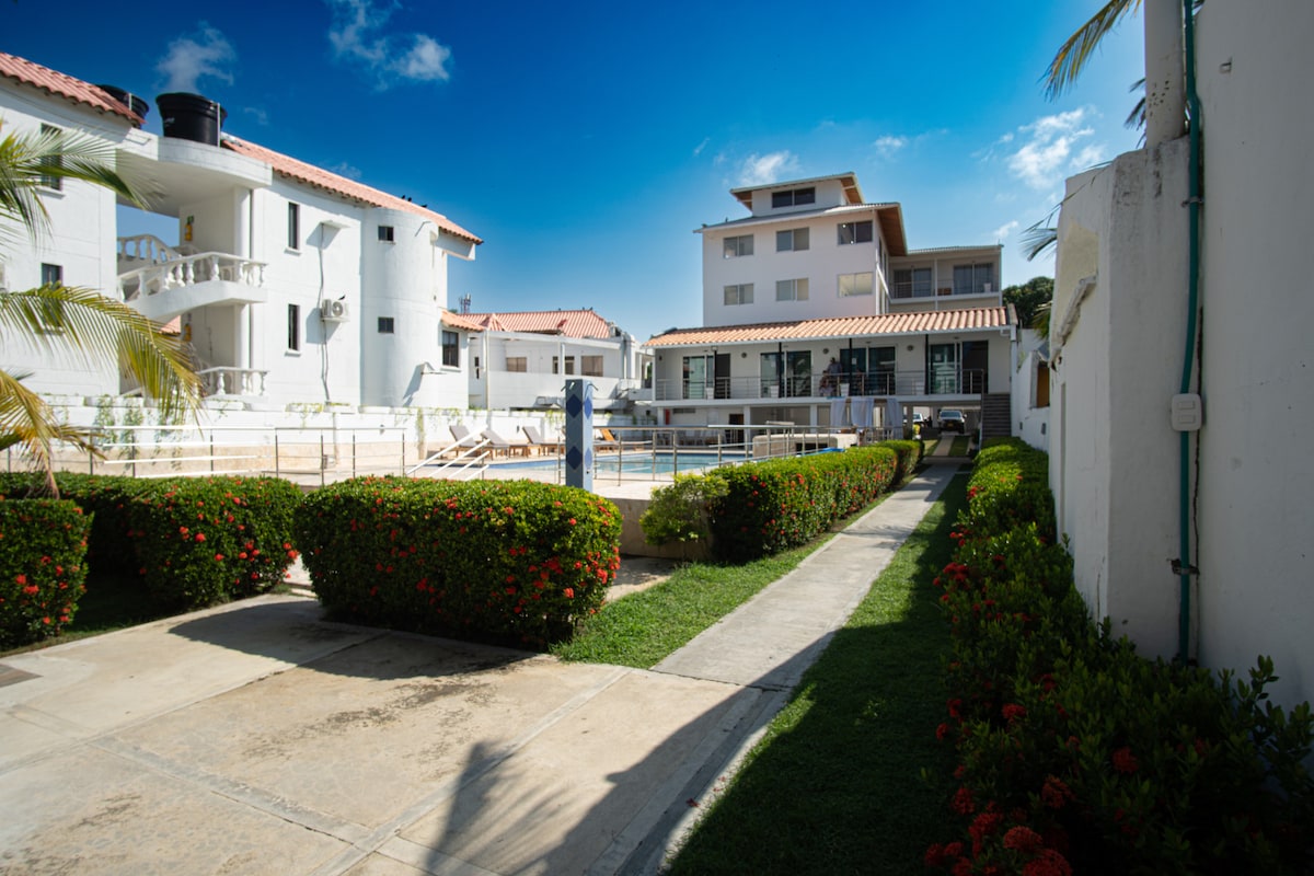 The outdoor area features well-maintained gardens with vibrant green hedges and blooming flowers. A clear blue sky is visible above, highlighting the modern buildings surrounding the spacious pool. The pool area is accessible, with lounge chairs and umbrellas positioned for relaxation.