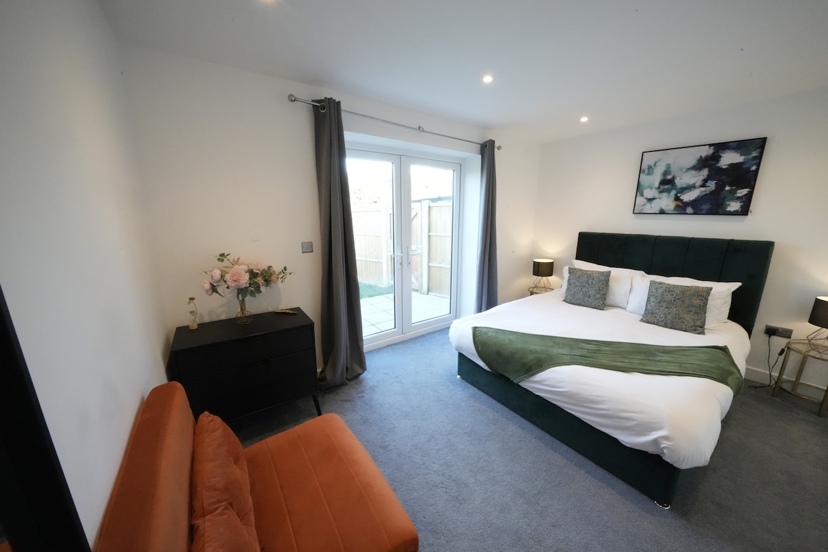 A well-lit bedroom features a spacious bed dressed in crisp white linens, accented with a green throw. A cozy seating area with an orange sofa is present, alongside a black dresser. Large glass doors lead to a private outdoor space, framed by grey curtains.
