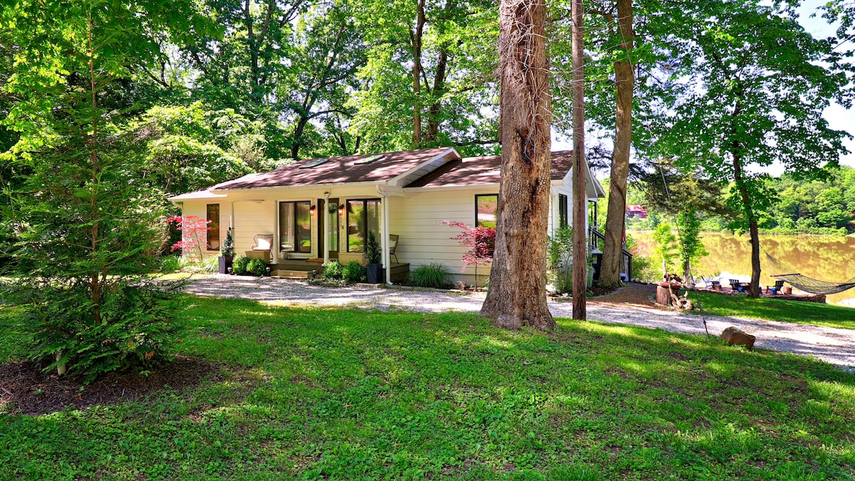 A single-story house is nestled among trees, surrounded by a well-maintained lawn. Natural light highlights the front porch, which features seating arrangements. A gravel path leads from the driveway to the entrance, with hints of the lake visible in the background.