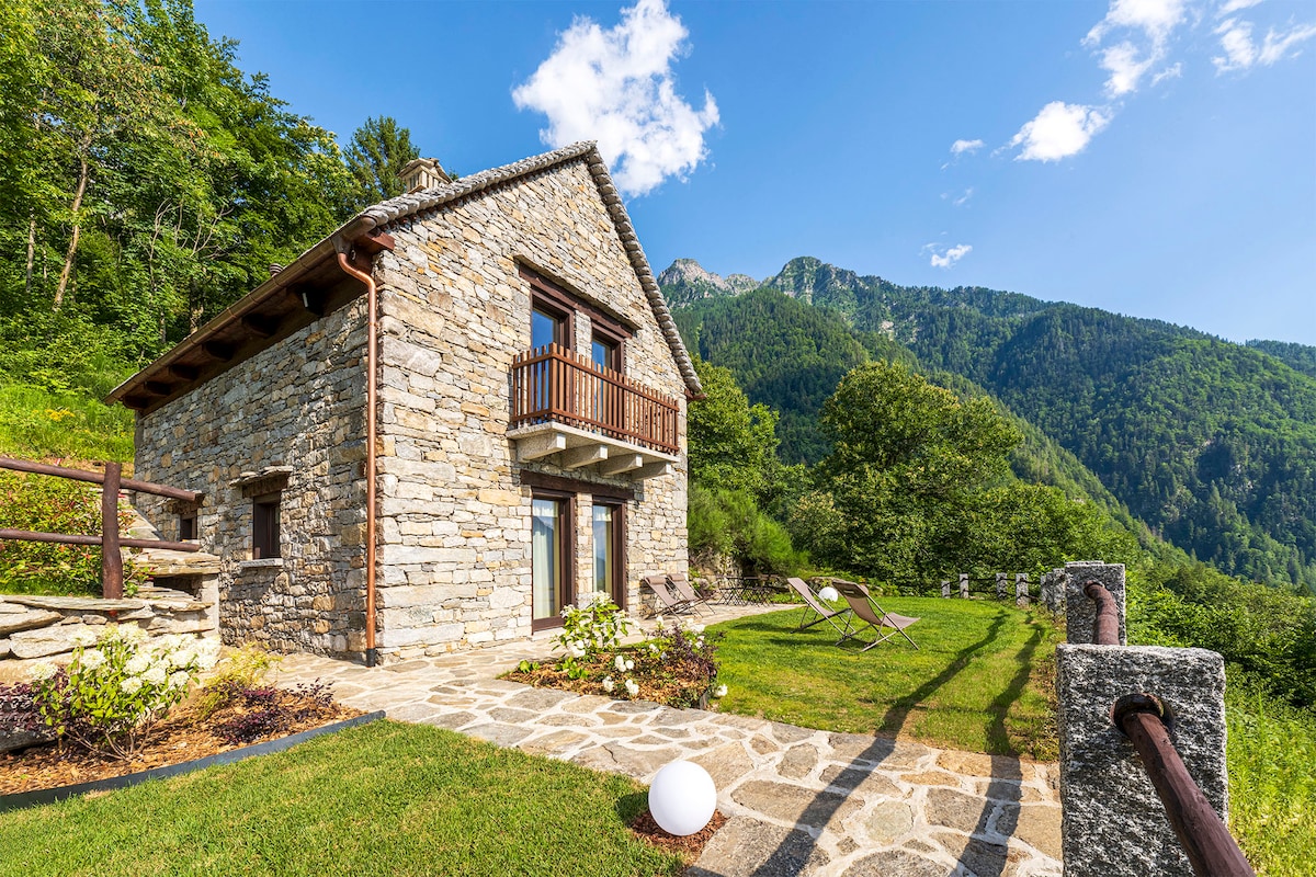 A charming stone cabin is surrounded by lush greenery, showcasing a well-maintained lawn and a stone pathway. A balcony extends from the upper floor, offering scenic views of the mountains. Sunlight bathes the cabin, highlighting its rustic architecture.