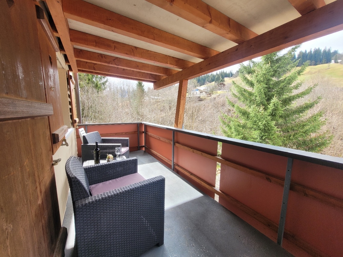 A balcony area is depicted with two dark wicker chairs and a small table, providing an outdoor space. The wooden beams overhead and railing add structural interest, while a view of trees and hills is visible in the background.