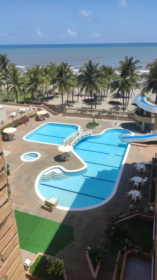 An expansive outdoor area features multiple pools, interspersed with sun loungers and tables. Lush palm trees line the backdrop, leading to a sandy beach and the shimmering sea beyond. Clear blue skies enhance the sunny setting, making it an inviting retreat for relaxation.