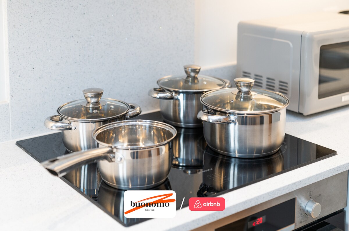 A sleek kitchen countertop features a modern stovetop with four stainless steel pots. Each pot is equipped with a glass lid, allowing visibility of the contents inside. A microwave sits nearby, completing the functional kitchen space.