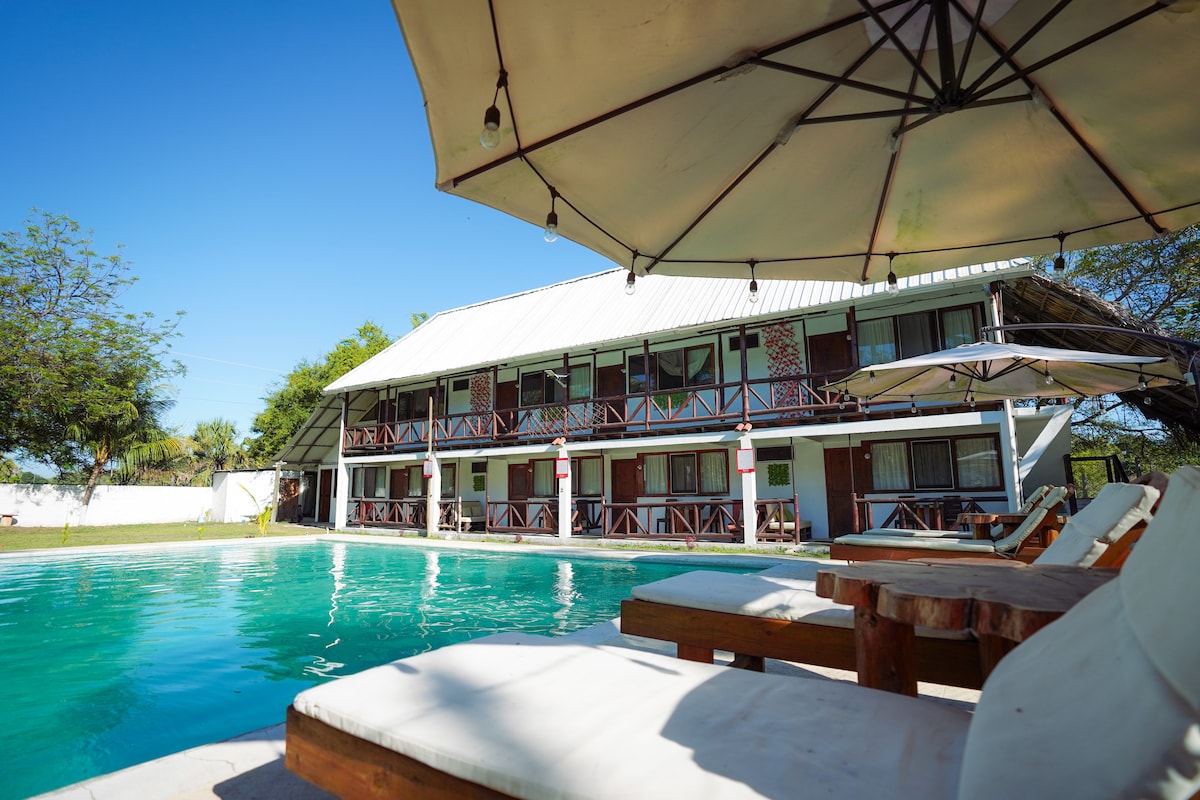 A swimming pool is featured in the foreground, bordered by wooden lounge chairs with cushions. In the background, a multi-level building displays a combination of balconies and large windows, all set against a clear blue sky and surrounded by greenery.