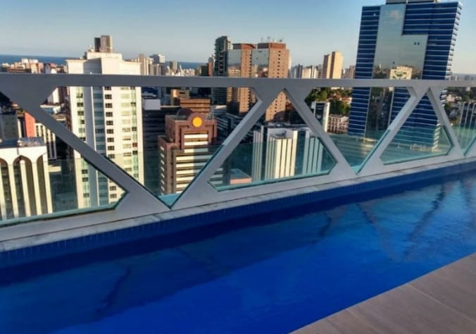 A rooftop terrace features a sleek pool with clear blue water, providing views of the surrounding cityscape. Modern high-rise buildings and a distant coastline are visible, creating a vibrant urban backdrop.