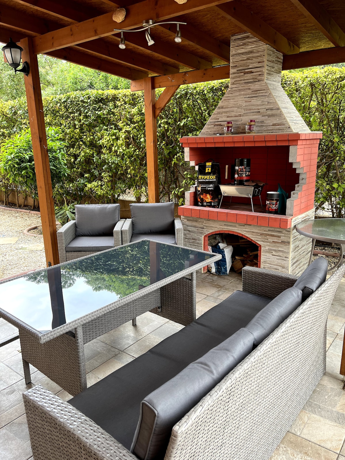 An outdoor seating area features a sturdy glass-topped table surrounded by comfortable wicker chairs. A built-in barbecue is positioned under a wooden gazebo, with stone and brick elements, enhancing the space for outdoor gatherings. Lush greenery provides a natural backdrop.