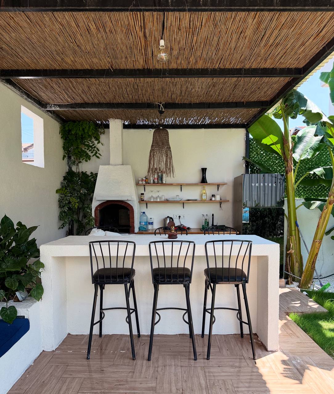 An outdoor bar area features three tall black stools arranged around a white counter. A chimney with a brick oven is seen in the background, accompanied by a shelf displaying various kitchen items. Green plants and a canopy provide shade and a natural ambience.
