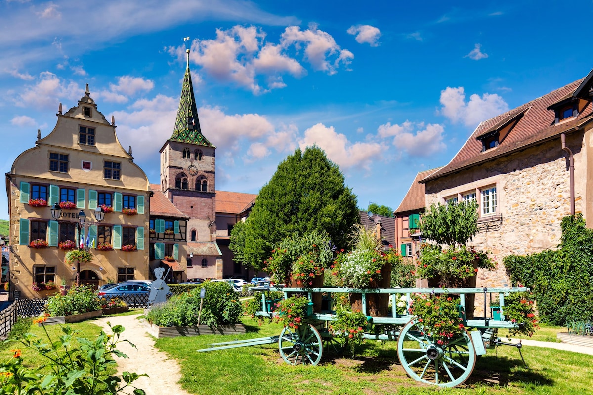 A vibrant village scene showcases well-preserved historic buildings with colorful shutters and flower boxes. A traditional cart adorned with blooming plants is positioned in a grassy area, while a tall church steeple and lush trees provide a picturesque backdrop under a blue sky with scattered clouds.