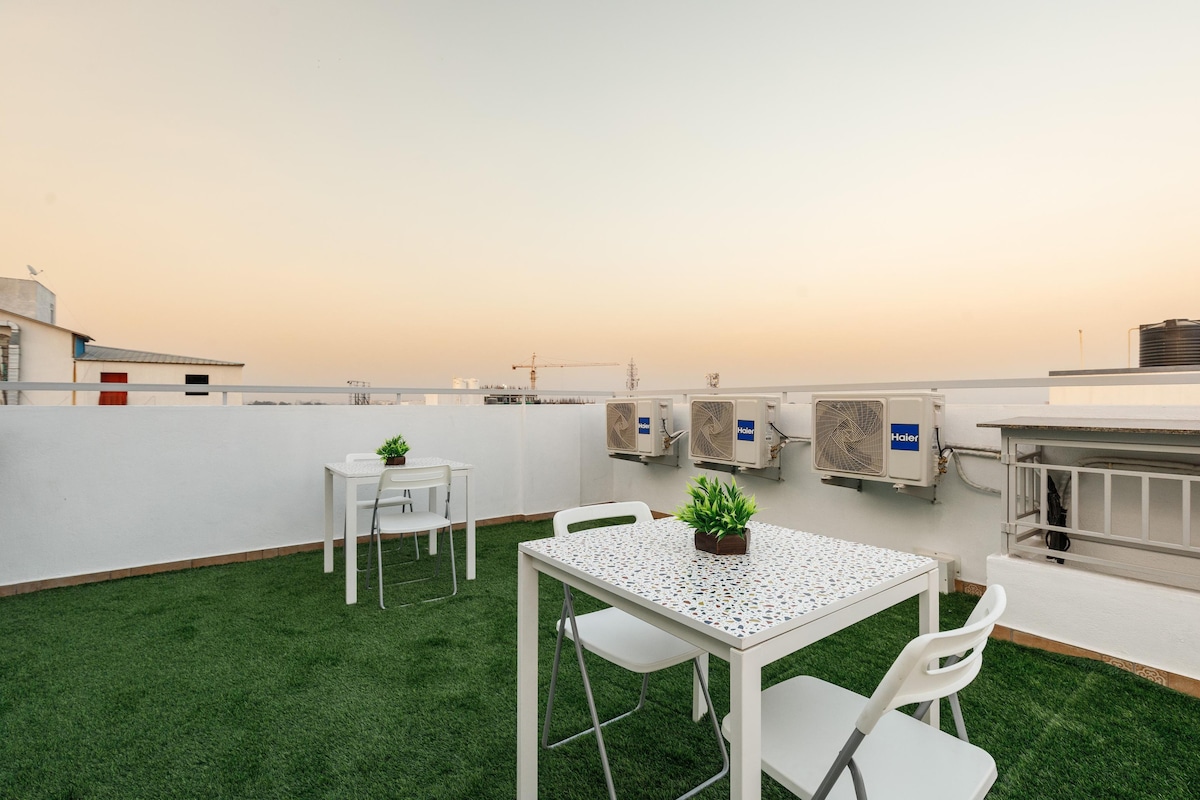 A terrace garden area is presented, featuring white outdoor tables and chairs set atop green turf. Potted plants add a touch of greenery, while air conditioning units are visible in the background against a soft sunset sky.