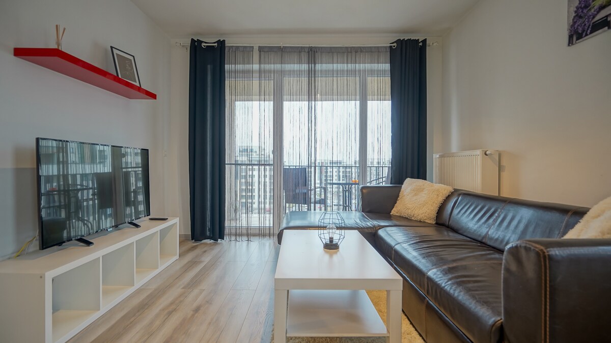 A modern living area features a comfortable leather sofa with soft pillows. A sleek white coffee table sits in front of it, while a television is mounted against the wall. Floor-to-ceiling curtains filter natural light from a balcony door, enhancing the open feel.