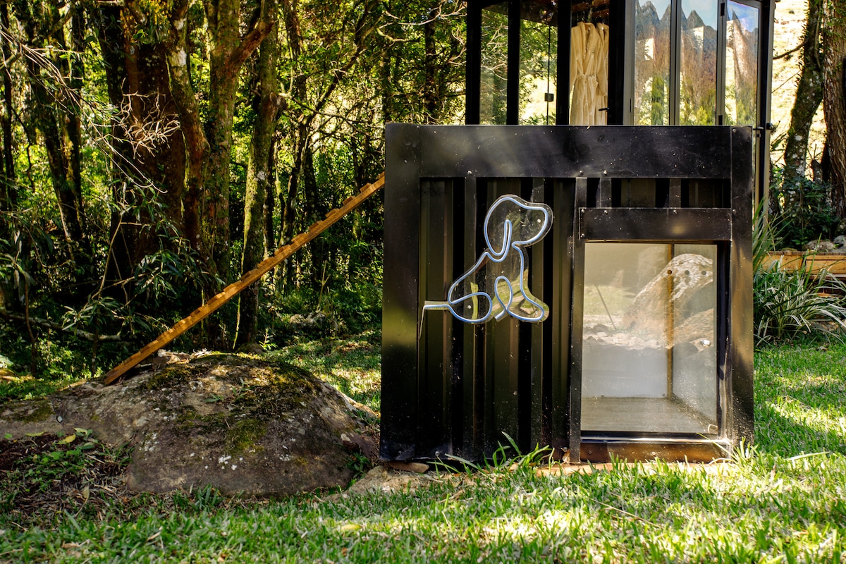 A pet house constructed from dark materials offers shelter, situated on a grassy area surrounded by trees. An illustration of a dog is displayed on the side, enhancing its inviting appearance. The entrance is designed for easy access, promoting safety for small to medium-sized pets.