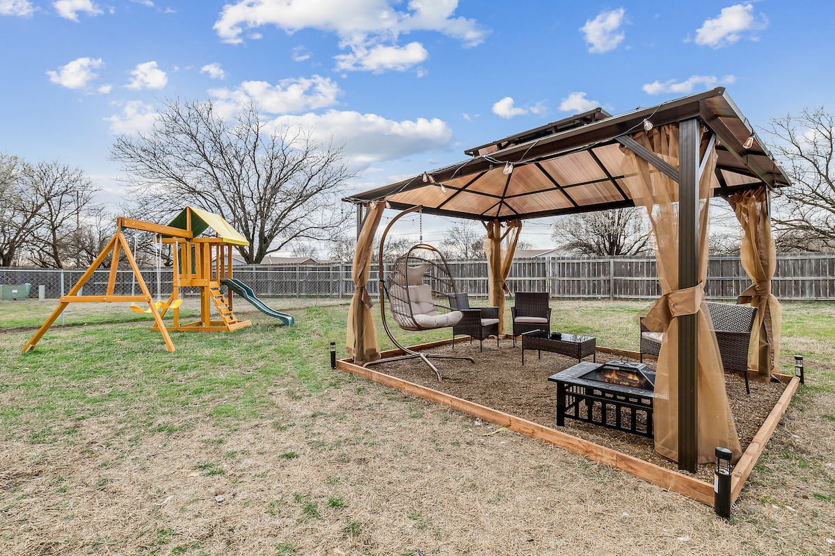 An outdoor space features a gazebo with draped fabric and a fire pit, accompanied by comfortable seating. Nearby, a colorful playground set invites children to play with swings and a slide, all set against a backdrop of spacious grassy areas and scattered trees.