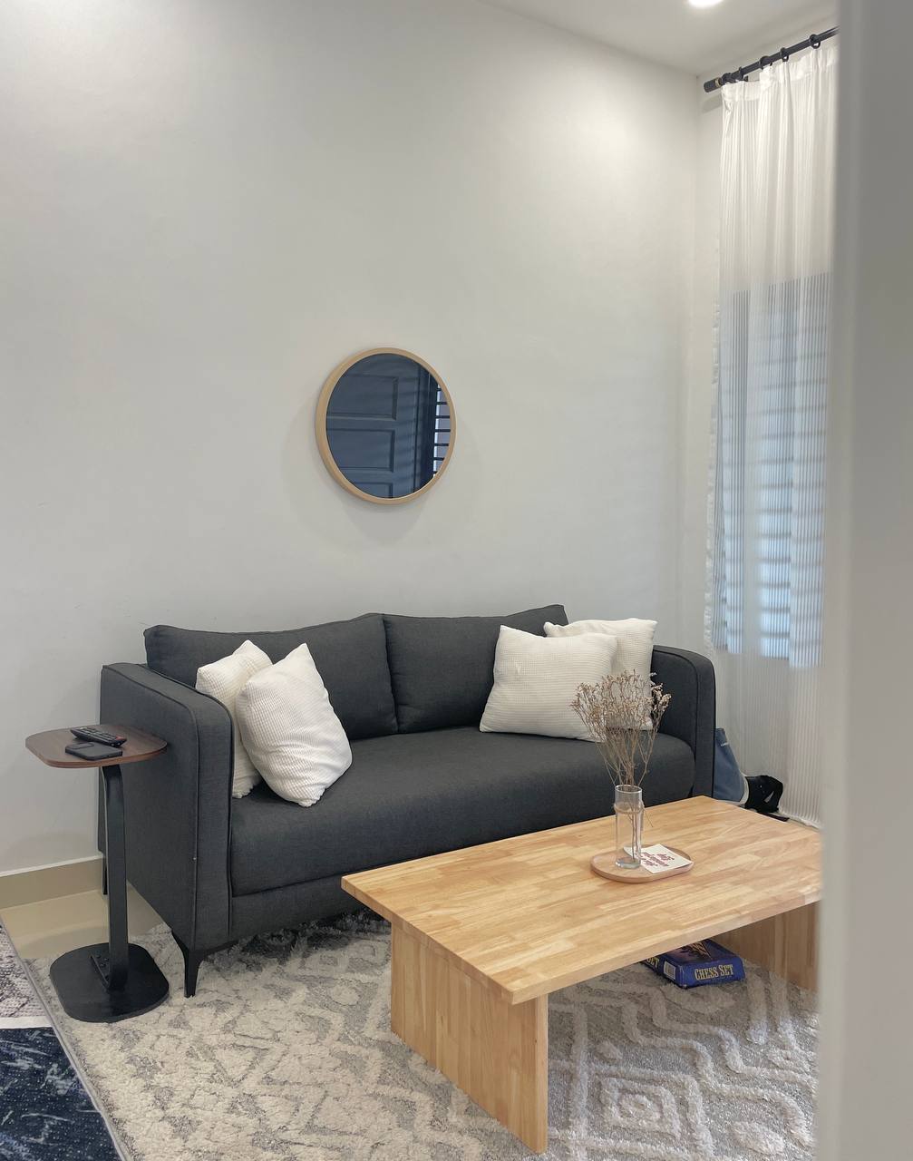 A cozy living area features a dark grey sofa adorned with four white cushions. A wooden coffee table is positioned in front, displaying a small vase with dried flowers. Natural light enters through the window, complemented by sheer curtains.