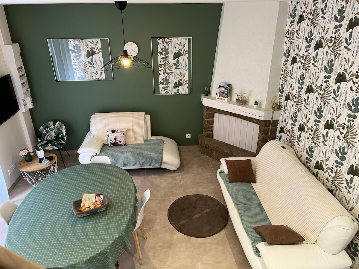 The living area features a green accent wall adorned with leaf-patterned wallpaper. A round table is set with chairs, while two sofas are arranged for comfort. A decorative clock hangs above, and natural light illuminates the space through large mirrors reflecting the furnishings.