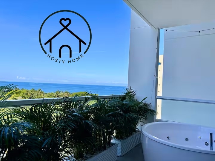 A private terrace is depicted with a jacuzzi surrounded by lush green plants. The expansive view of the ocean is visible in the background, with a clear blue sky creating a serene atmosphere.