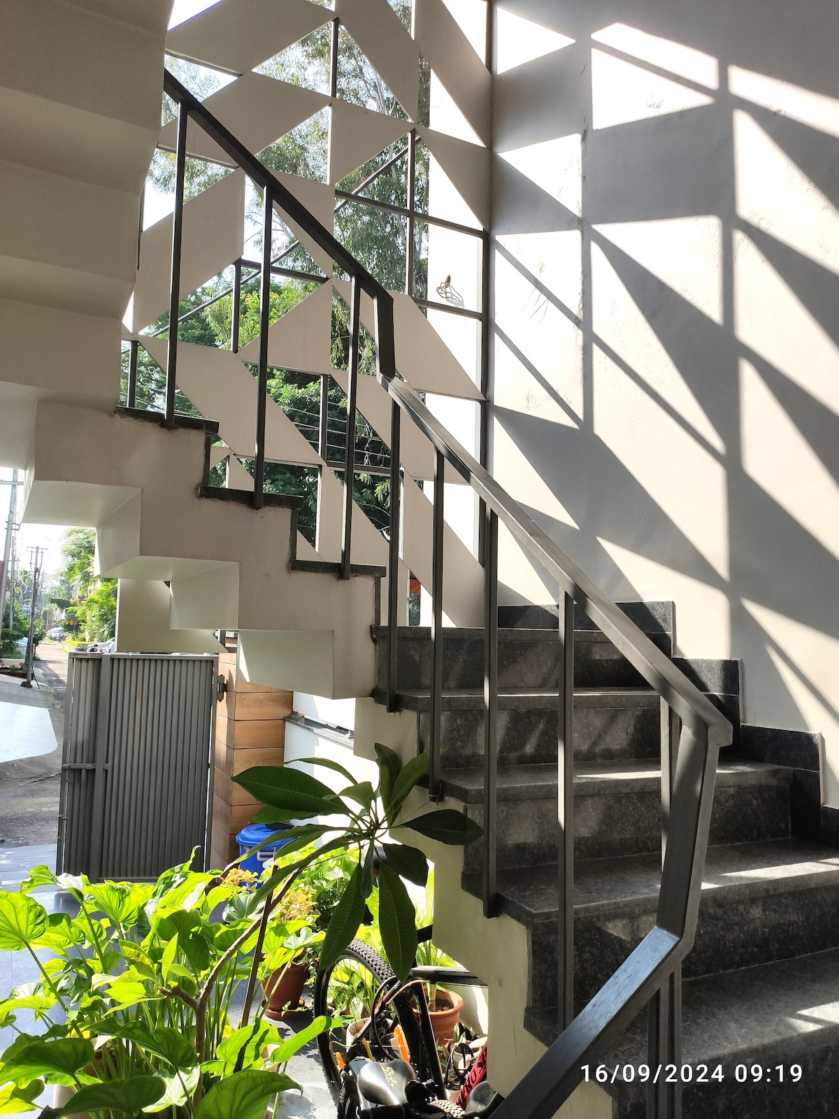 A staircase is illuminated by natural light, showcasing a modern design with angular windows. A metal railing provides support, while greenery at the base enhances the inviting ambiance of the space.