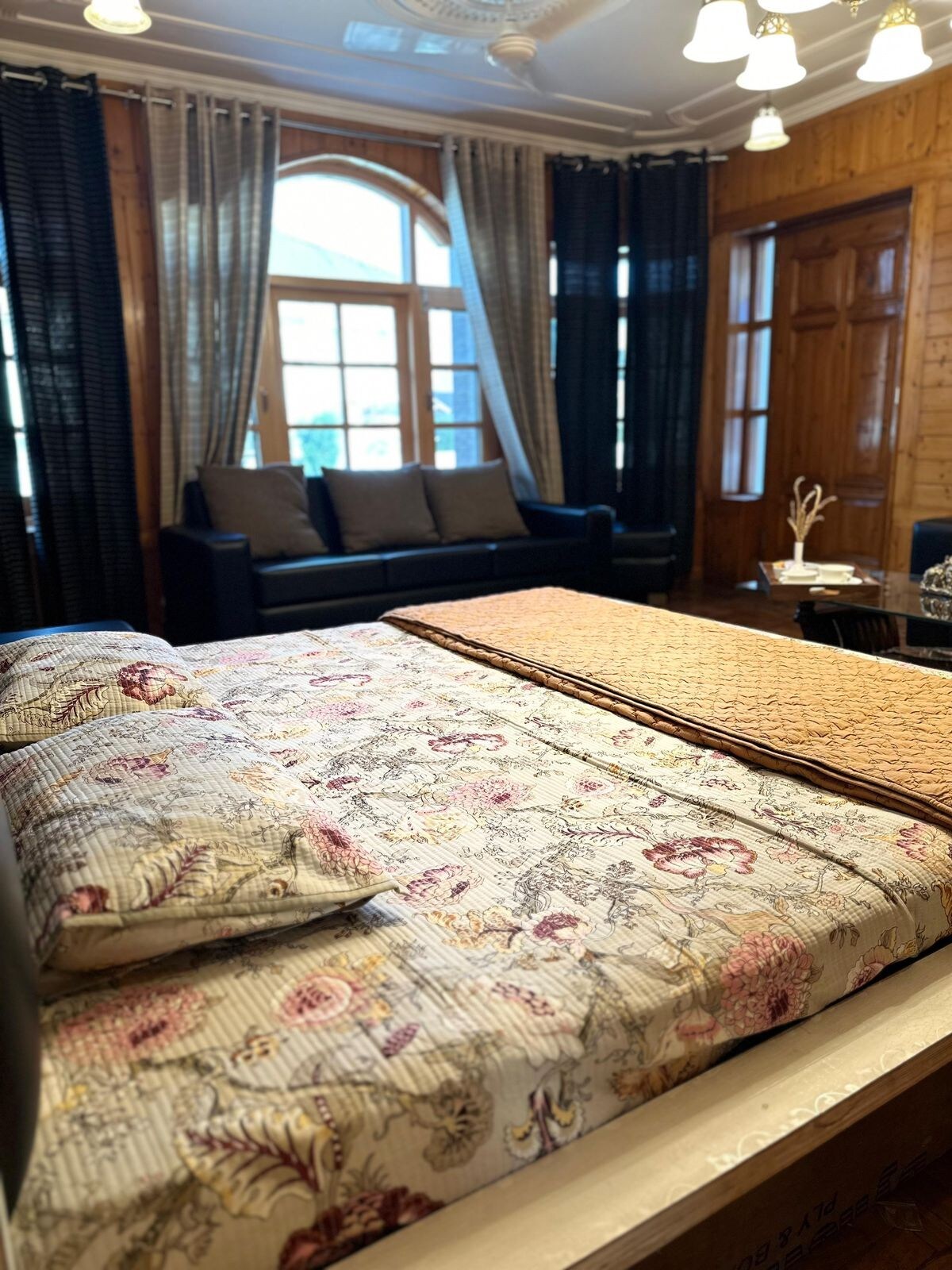 A cozy sleeping area features a patterned bedding arrangement atop a spacious bed. Soft textures are complemented by elegant wooden paneling and large windows dressed with light curtains. A seating area with a dark sofa is visible in the background.