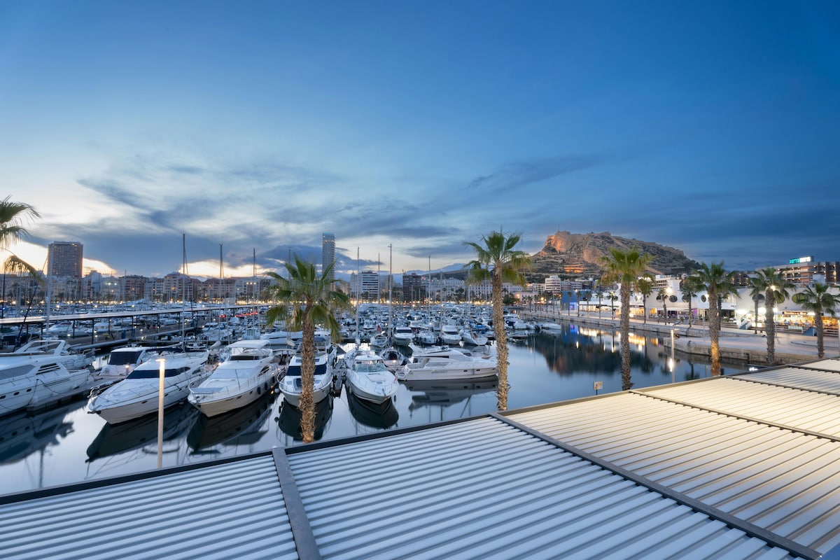 A tranquil view of a marina is presented, featuring numerous boats gently bobbing in the water. Palm trees line the waterfront, framed by a vibrant sky transitioning from blue to twilight. Reflections of the boats and lights create a serene atmosphere.