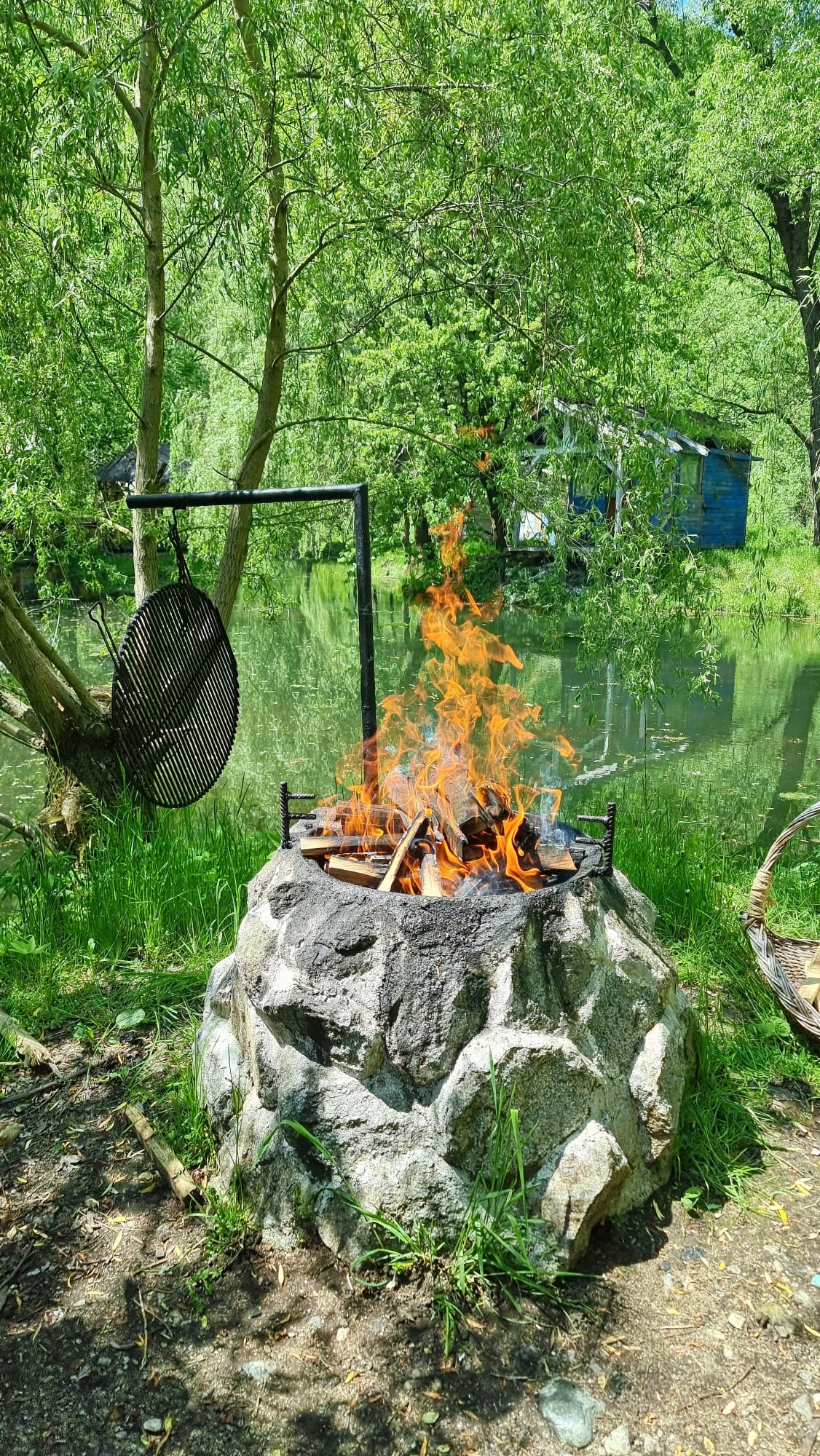 A fire pit made of stone is visible, with vibrant flames rising from burning wood. Set against a lush green landscape, the area is surrounded by trees, and a serene body of water is seen in the background.