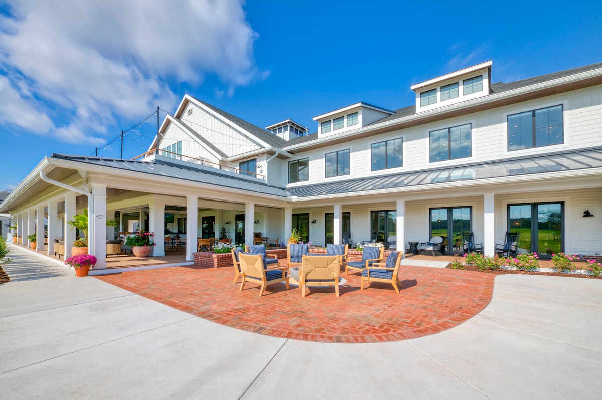 An outdoor area features a brick patio surrounded by comfortable seating, ideal for relaxation. The three-story home in the background showcases large windows and a welcoming facade, with lush landscaping enhancing the space. Clear skies provide a bright backdrop to the scene.