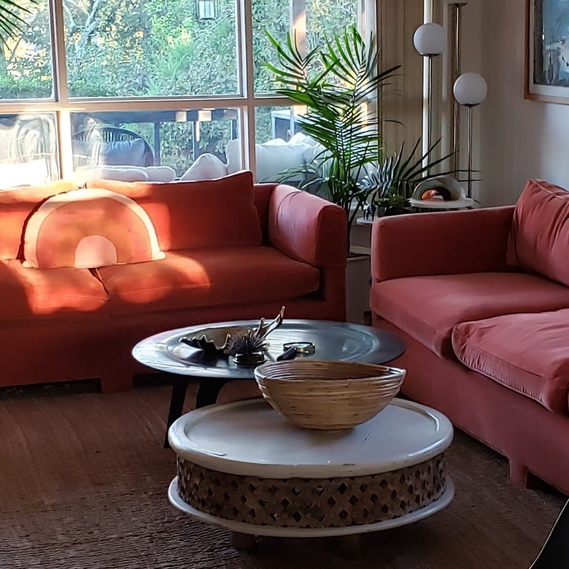 A bright and airy living space features large windows allowing natural light to fill the room. Two comfortable red sofas are arranged around a round coffee table, which holds a woven basket. Decorative plants add life to the cozy atmosphere.
